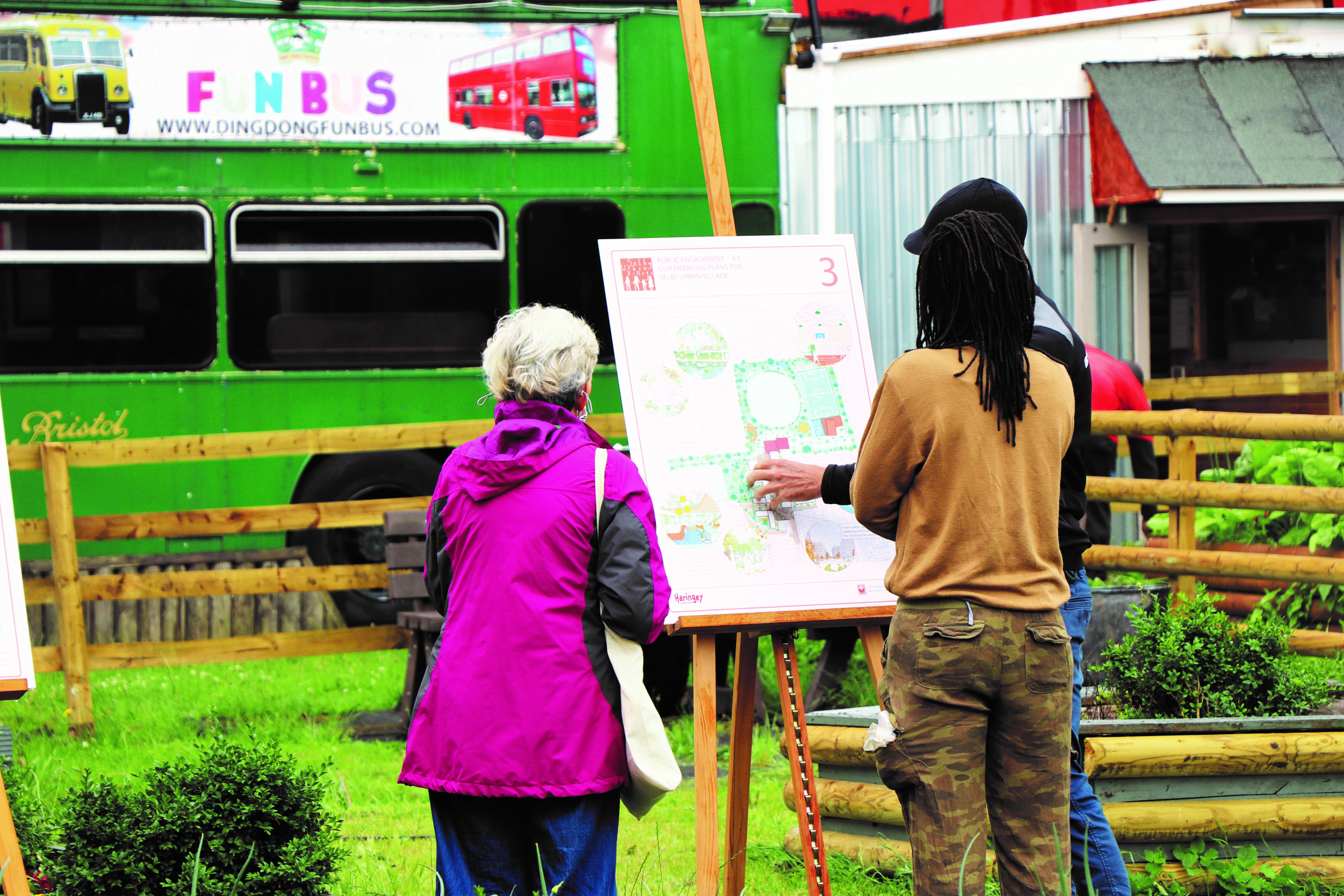 A map in a black cap, and black jumper and blue trousers, largely obscured from view, shows a woman with white hair and a pink coat and jeans and a woman with dreadlocks in a caramel top and camouflage trousers a map on a wooden easel. Behind them is a section of enclosed parkland and behind that is a green bus.