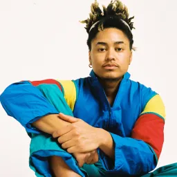 A picture of the artist Khaleb Brooks. He is wearing a brightly-coloured jacket, and sitting with his hands clasped over his right knee. He is mixed-race and has a single earring in his right ear.