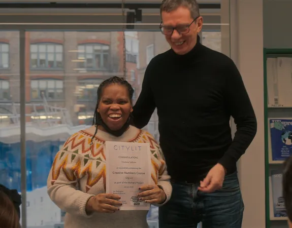 A young person with a huge grin holds up their certificate. Their tutor stands next to them smiling too, while the rest of the class looks on.