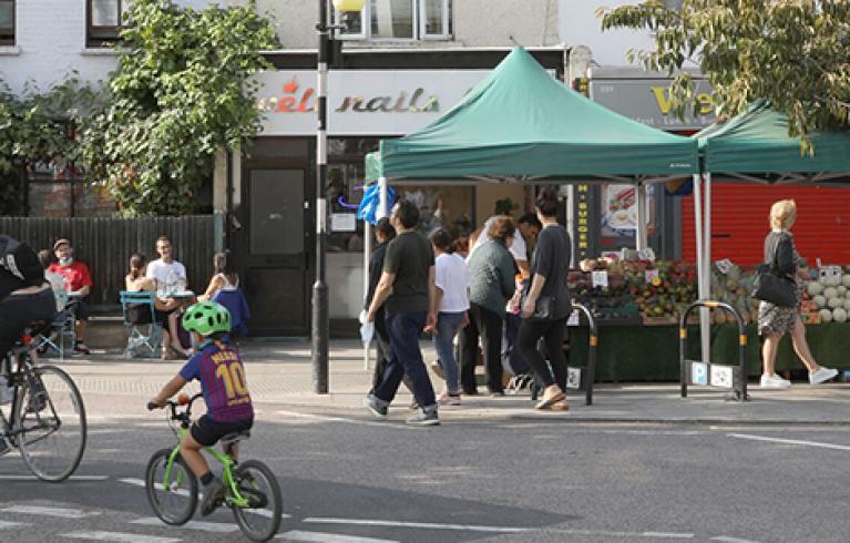 A high street in East London showing shops, market stalls, pedestrians who are sitting, cycling and walking.