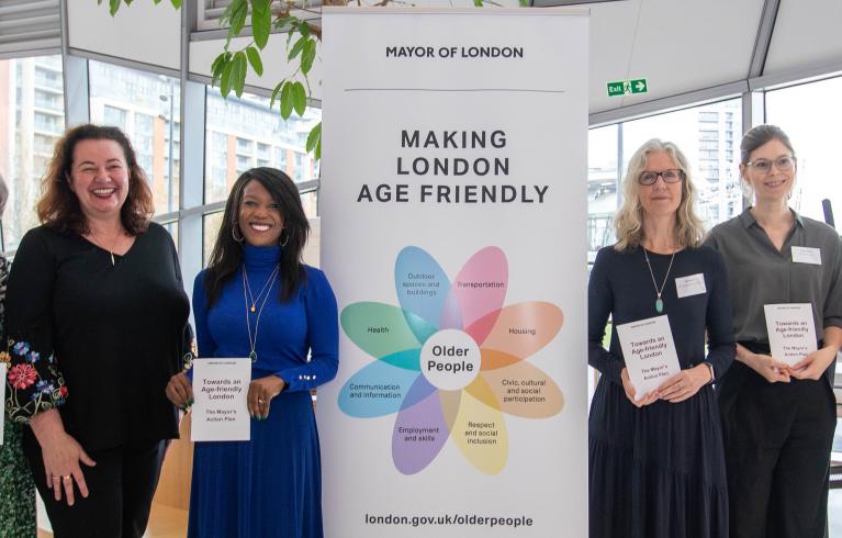 Six people of different ages stand next to a large banner at action plan launch event. The banner reads "Making London age friendly". 