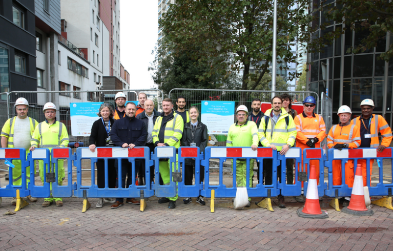 Large group of people from utility company's street service. They wear high vis clothing and are standing behind a temporary barrier on a city street.