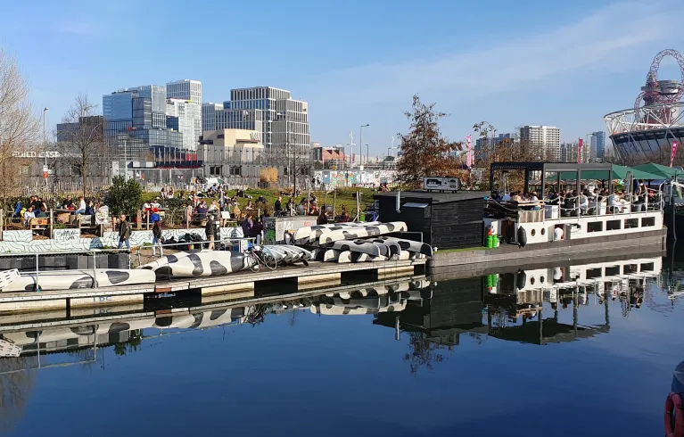 View across the canal at Hackney Wick. People sitting at an outdoor bar area overlooking the Queen Elizabeth Olympic Park and East Bank