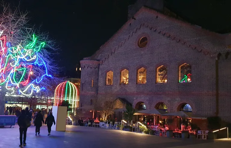 View at night across a square of a tree with neon lights, a birdcage light art installation and restaurant with outdoor seating