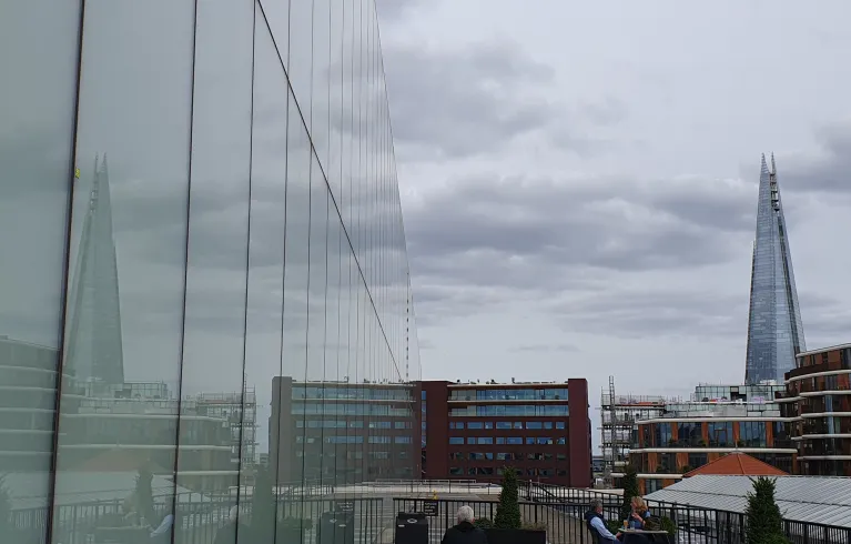 Side of a large glass building reflecting the view on a cloudy day of mixed architecture and the Shard in the distance.