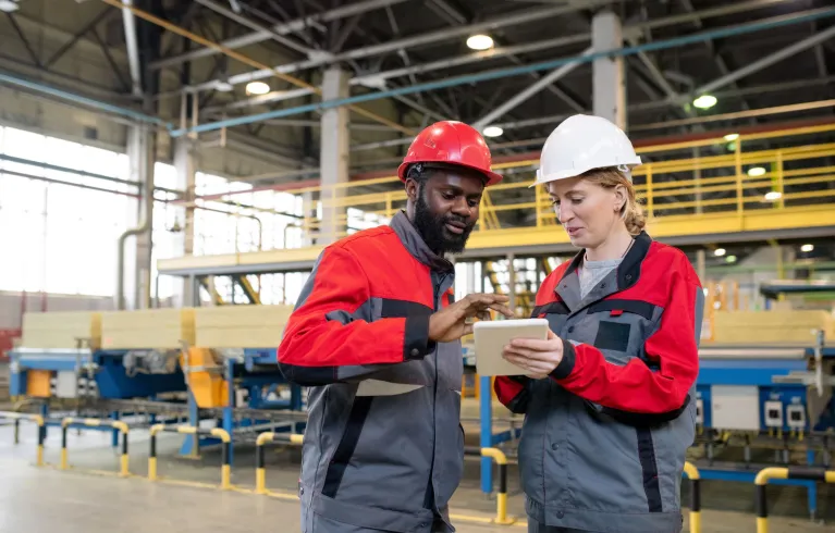 Two people wearing work uniform and helmet discussing information on a tablet at factory