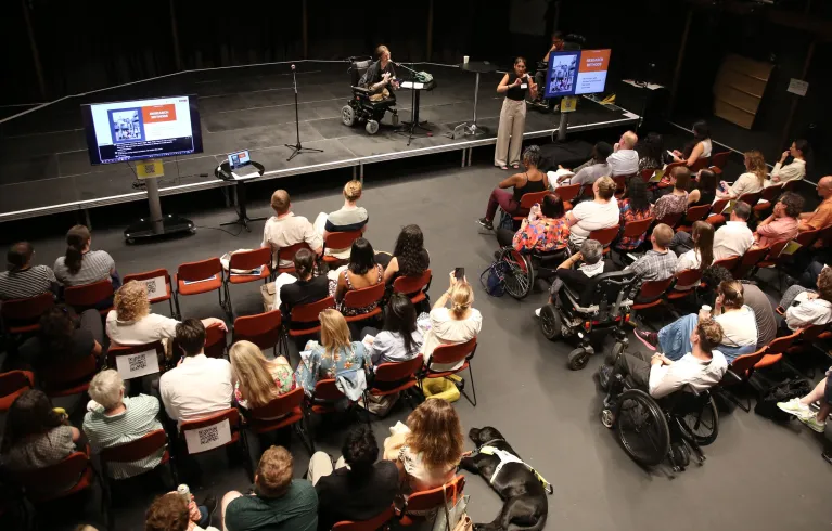 View of stage and audience from up in the gallery. Rows of chairs and wheelchairs line the audience, with a guide dog lying at the end of a row.  One speaker addresses the audience on stage, while a BSL interpreter signs.