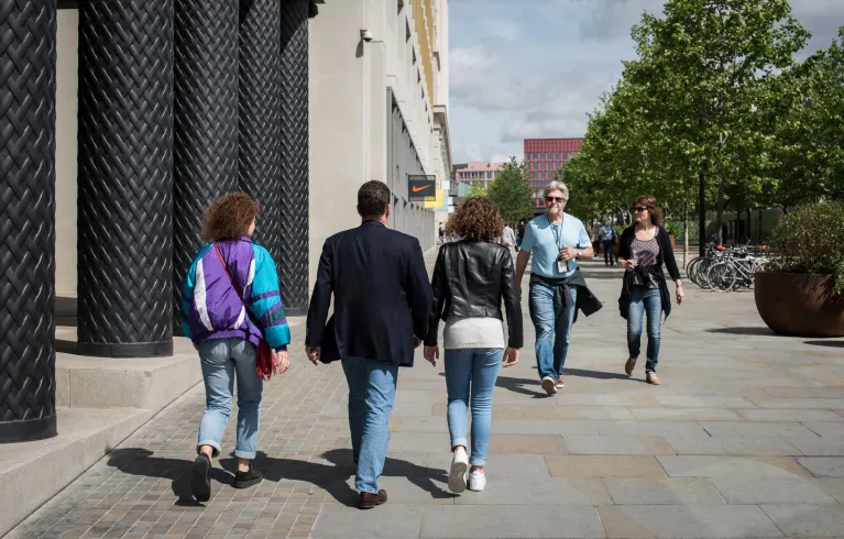 Three pedestrians walking with their backs towards the camera, while two others walk towards the camera. They are on a wide pavement with a columned building to the left of the image and a tree at the back right of the image