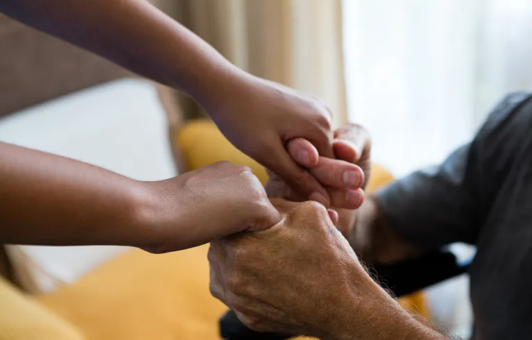 A carer supports an older person, holding their hands.