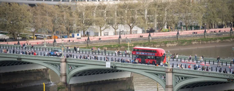 A red bus driving over Westminster bridge with the Thames river, members of the public and trees visible