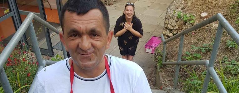 a man looks to camera holding two full plates of spaghetti and veggies, a woman stands behind him smiling with hands to her heart in a compassionate motion.