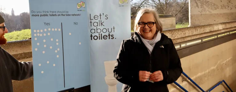 Caroline Russell poses with a smile next to a banner with lots of yes votes for more public toilets on the tube network.