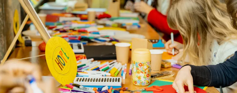 Close up of a busy table top with coloured pens and arts and crafts tools. There are people around the table, including a child drawing, and a sign at the forefront reads ‘hope’