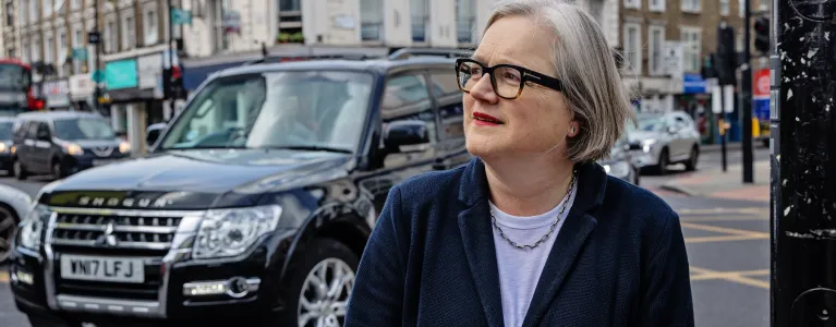 Caroline Russell, standing in front of moving traffic at a London street corner, looks pensively off to the lefthand side of the frame without smiling.