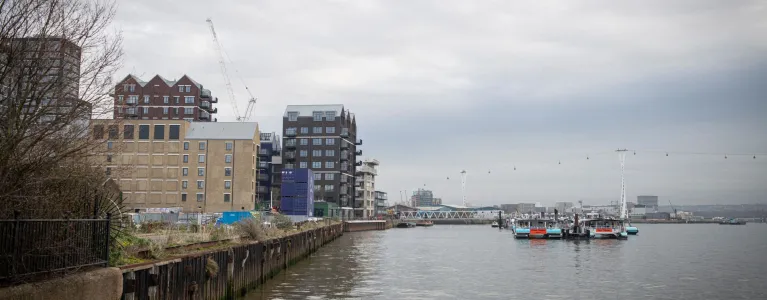 Wide angle image of the Thames River, looking towards the Thames Barrier. There are several buildings next to the river on the left-hand side of the image. The photo was taken on a cloudy, overcast day