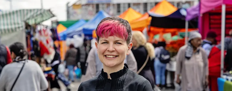 Zoë Garbett stands on a sunny day at Ridley Road Market in London. She is smiling straight at the camera wearing a black longsleeve blouse.