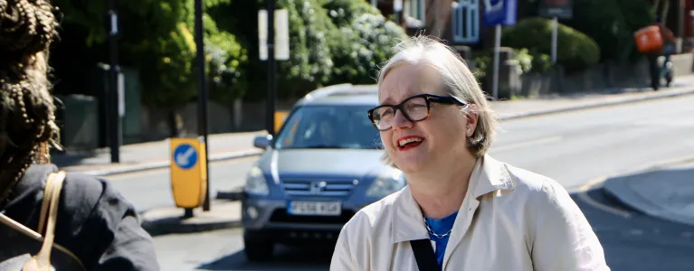 Caroline Russell campaigning for more push buttons on street crossings across London. She is smiling, wearing a white jacket and looking off to the left of the camera engaging with a Londoner. A car is visible in the background. 