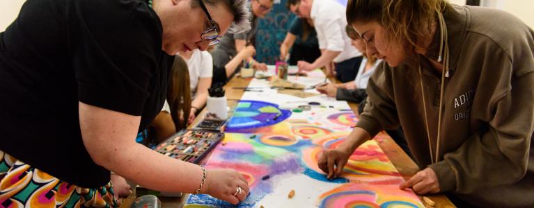 A group of people stand over a table, drawing with coloured chalks on a collaborative artwork which shows shapes and circles coming together