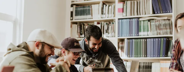 A group of people chat and laugh sat around a table with laptops in a library