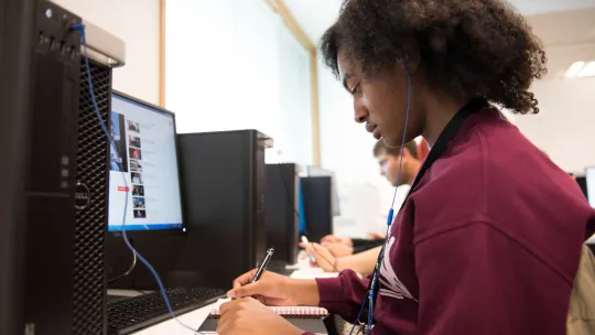 Young person sitting at a desk and writing down information found on a computer
