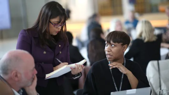 two women at a conference, one sitting down and talking to the other, who is writing in a notebook