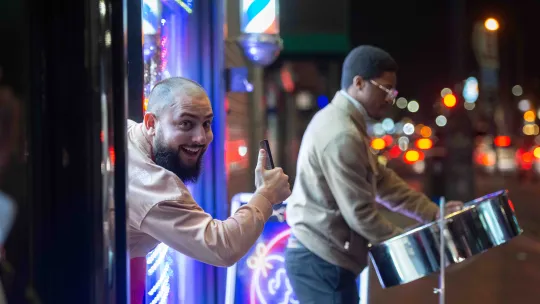 A steel pan performer and a man leaning out of a shop door sticking up his thumb in approval 