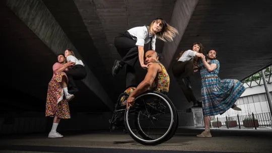 Three couples dancing under an underpass. The men wear dresses, while the women are jumping in formal wear. The dancer in the centre is sat in a wheelchair.