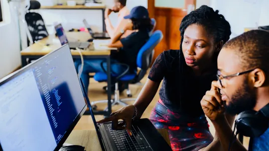 A woman and man look at a screen displaying software coding in an office. 