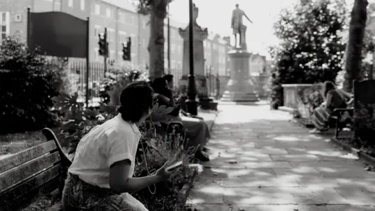A woman sat in a park in Bow, with headphones in, listening to audio story about the Gladstone statue which is in the background