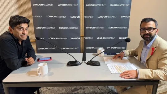 Two men with microphones looking at the camera with the London Assembly banner behind them.