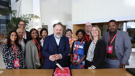 Group of people surrounding the Deputy Mayor for Business Howard Dawber cutting a pink Good Work Standard cake