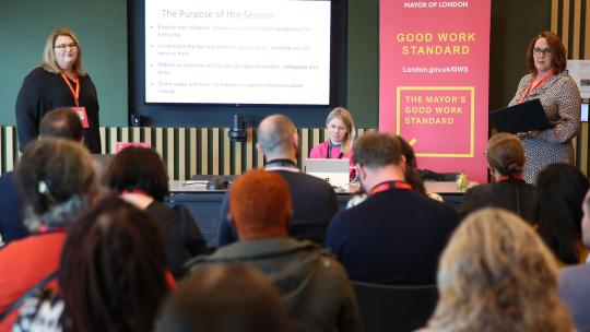 2 presenters Katie Coates and Hazel Leighfield presenting at the front of a Committee Room with an audience. A pink Good Work Standard banner is in the background