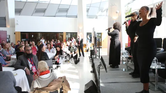 3 singers singing on stage at City Hall to a large audience in London Living Room