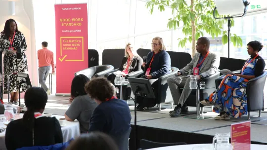 Panel of 4 people sitting on stage in armchairs in London Living Room with Good Work Standard pink banner behind them