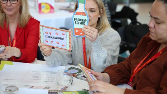 3 people at a table looking closely at colourful printed shapes in the shape of a bottle as part of workshop