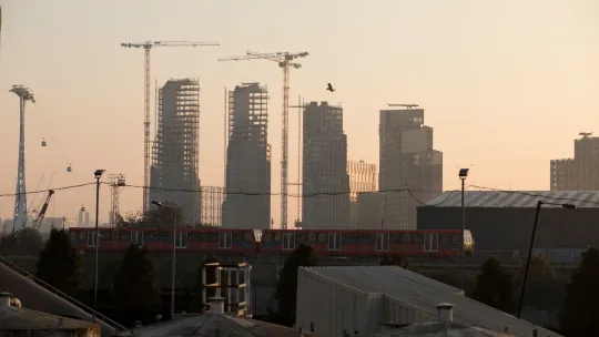 Construction site of 4 tower blocks at dusk. DLR train passing in front, cable car overhead lines to the left.