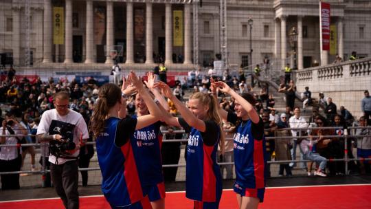 Four female players in blue and red Junior NBA 3v3 uniforms celebrate with high-fives on a basketball court in Trafalgar Square, surrounded by spectators.