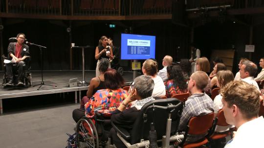 The audience look toward the stage where James Lee is welcoming everyone to the event. There is a screen to the right of the stage detailing the day's agenda.