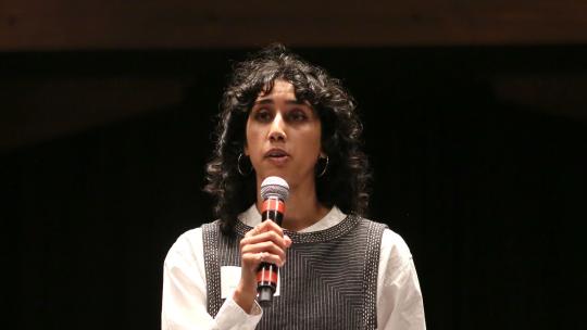 Close up of Ash Roa stood on stage standing out against a completely black background as she addresses the audience. She is wearing white trousers, a white shirt and grey vest. 