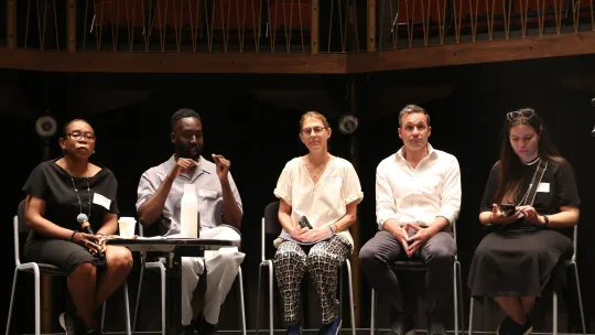 Five case study speakers sit on stalls on stage. The second person along signs to the audience while the others listen to the BSL interpreter.