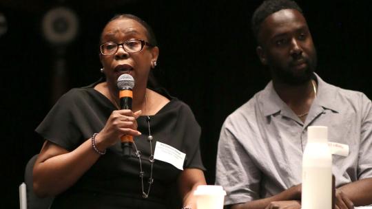 A black woman sits on stage in a black dress, holding a microphone and addressing the audience.