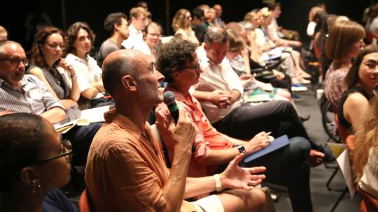 An audience member asks a question. He is wearing an orange shirt and white shorts and gestures with his left hand while speaking into the mic in his right.