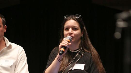 A white woman in a long black dress sits on a stall on stage and speaks into a handheld microphone.
