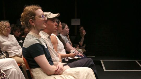 A woman and her guide dog sit at the end of the front row looking up at the stage.