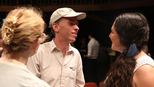 Three people are standing in conversation smiling with one another. A man in a white shirt and white cap stands in the middle, while two women are listening either side, with their bodies turned towards him.