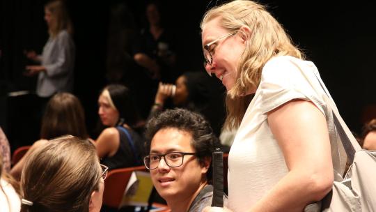 A woman in a white t-shirt holding a cane stands, smiles while in conversation with two individuals sat down.