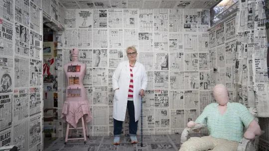 A woman in a long white coat, blue turned up jeans, pale blue trainers with red tips, and a red and white striped top, stands with a blue walking stick towards the back of a room. She has short blond hair, dark framed glasses and red lipstick. The room is covered - walls and ceiling with newspaper. There is a soft stuffed mannequin sat in an armchair, also covered in newspaper, at the front right. They wear a pale green shirt and light trousers. In the back left is a pink tailor dummy with a teapot. 