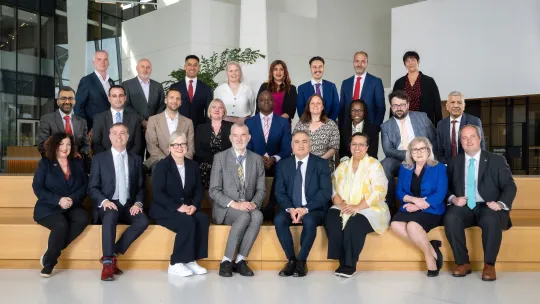 Diverse team of professionals poses in three rows on modern office stairs, wearing business attire and smiling in a welcoming group portrait.