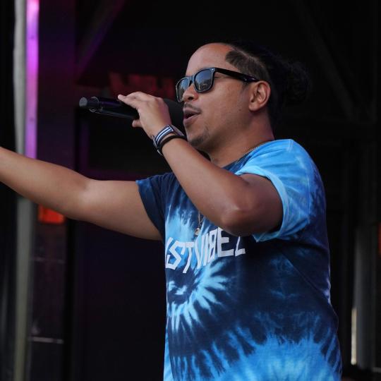 A male singer wearing sunglasses and a blue t-shirt preforming with a microphone
