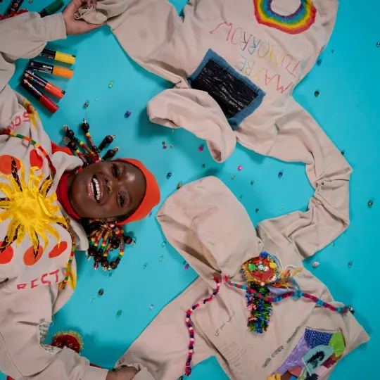 A young person with colourful braids lies on the floor looking straight up at the camera with a wide smile, wearing a hoodie with a sun that says ‘OK’ and holding two other hoodies with colourful writing and beads spread on the floor. It is vibrant and colourful.